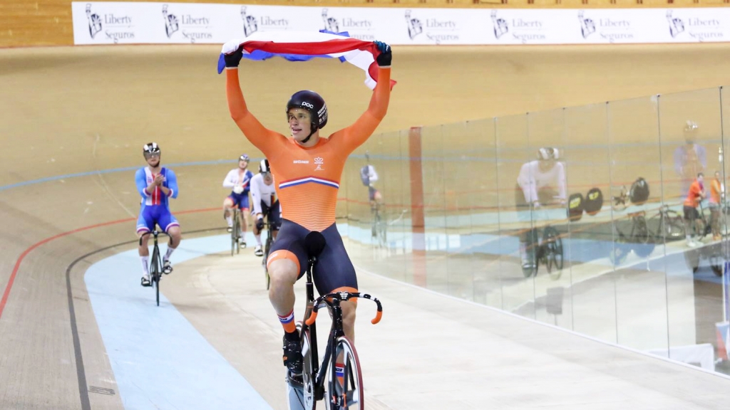 Nederlandse baanrenner juicht met de vlag boven zijn hoofd na winst op het EK baan U23, met achtervolgers in de baan