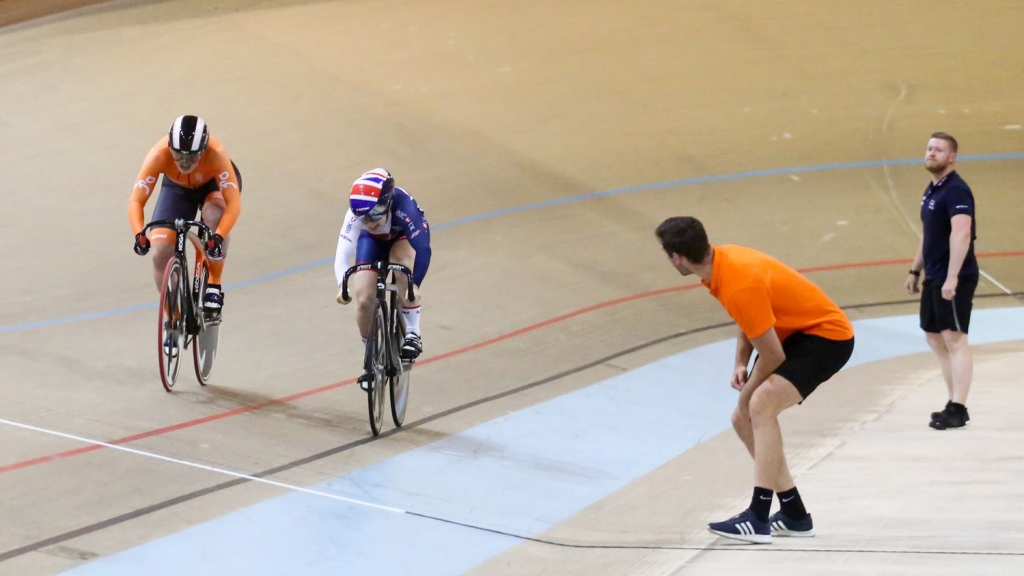Nederlandse baanrenner in oranje sprint zij aan zij met een Britse renner op de wielerbaan, terwijl coaches toekijken