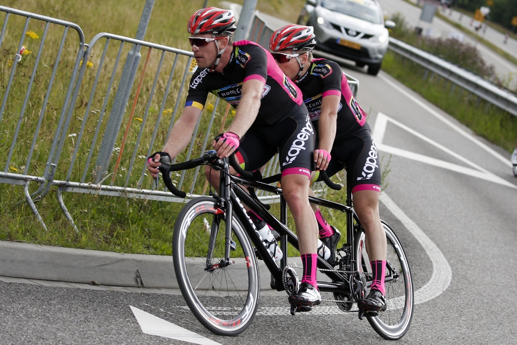 Tristan Bangma en zijn piloot rijden op een tandem in roze-zwarte tenues over de weg tijdens een para-cyclingwedstrijd